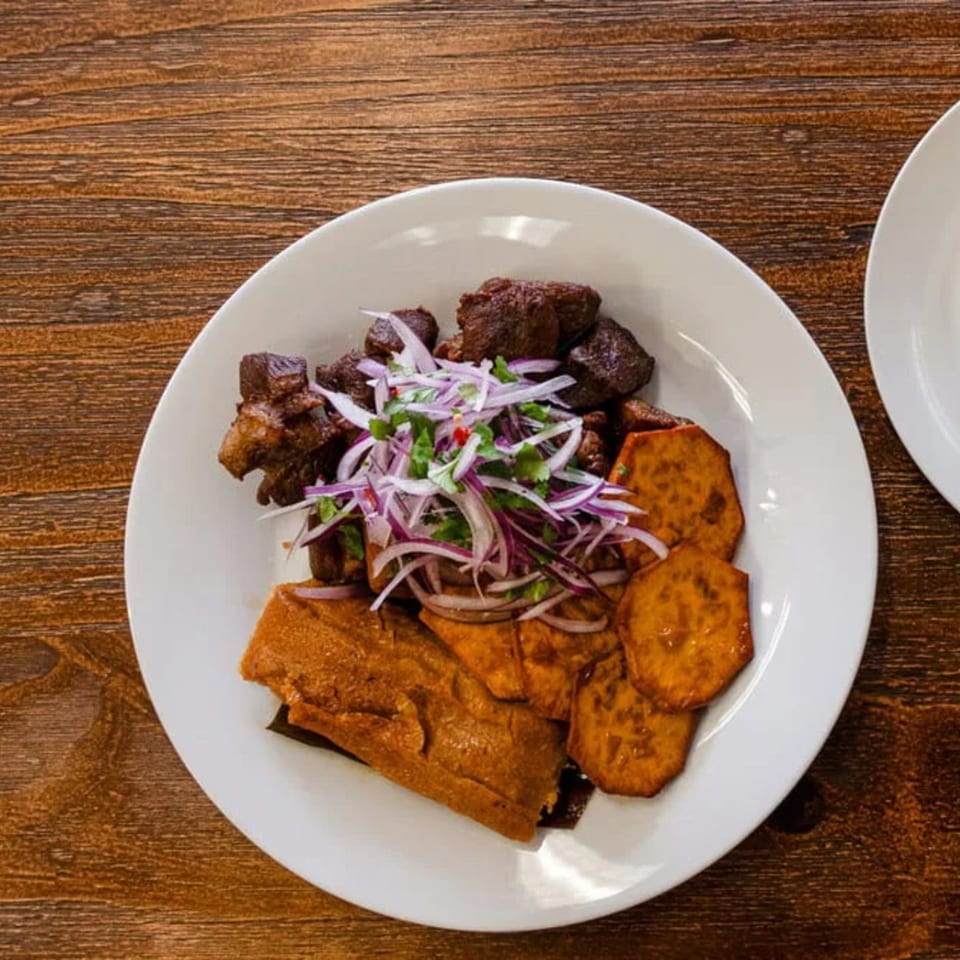 Peruvian dish with beef, sweet potato slices, and a tamale, garnished with red onions and cilantro.