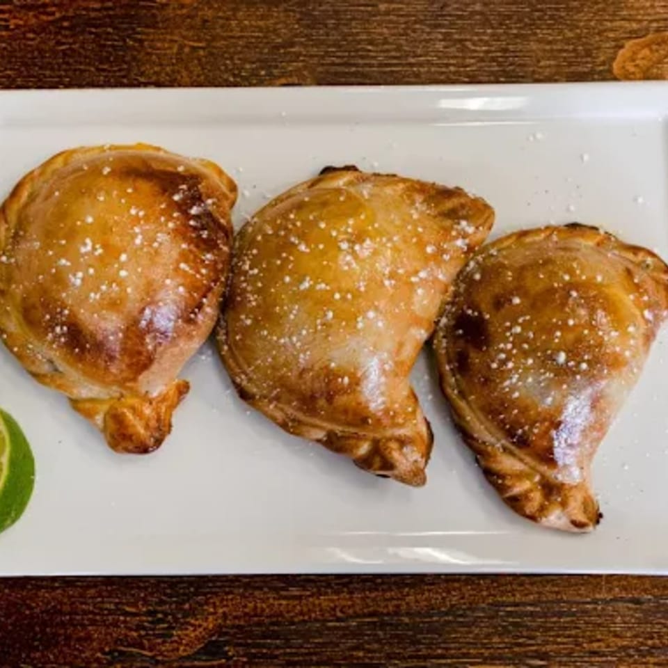 Three golden-brown Peruvian empanadas dusted with powdered sugar and a lime wedge on a white plate.