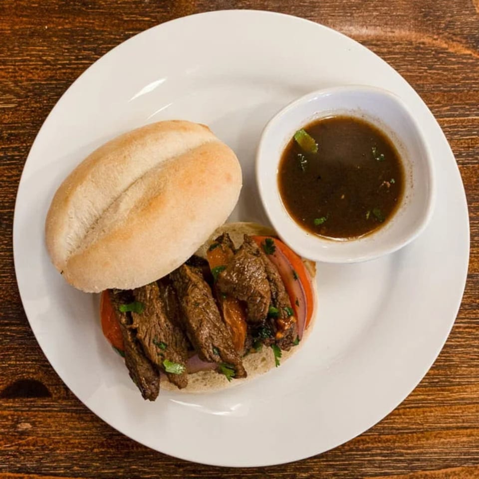 Peruvian Lomo Saltado sandwich with steak, tomatoes, onions, and cilantro, served with a side of au jus.