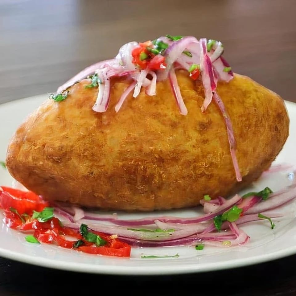 A golden-brown Peruvian empanada, topped with red onions, diced tomatoes, and green herbs, on a white plate.